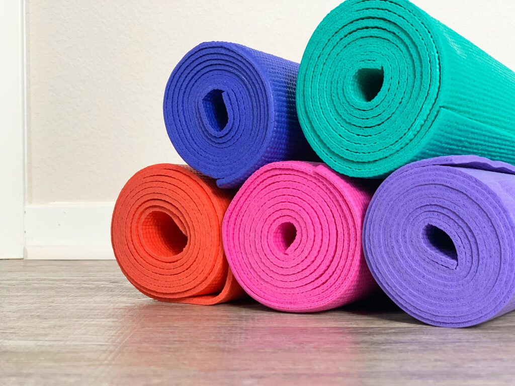 unclose image of yoga mats rolled up on the floor. Yoga mats are colorful. Featuring orange, pink, purple, indigo, and teal.