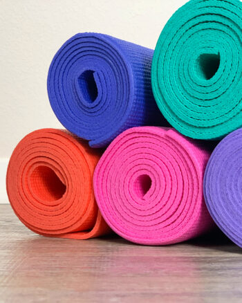 unclose image of yoga mats rolled up on the floor. Yoga mats are colorful. Featuring orange, pink, purple, indigo, and teal.